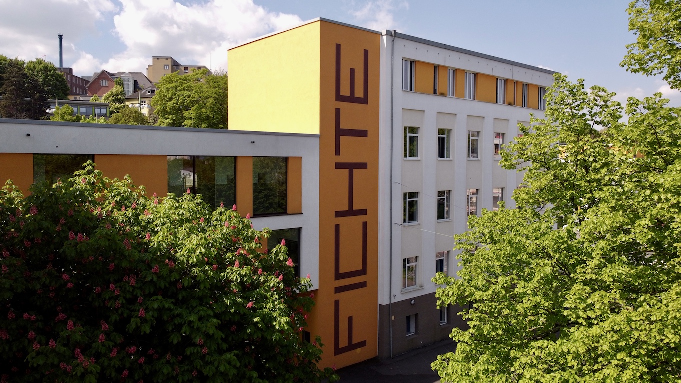 Weißes Schulgebäude mit hellorangenen Farbakzenten und riesigem vertikalem Fichte-Schriftzug. Drumherum Bäume.